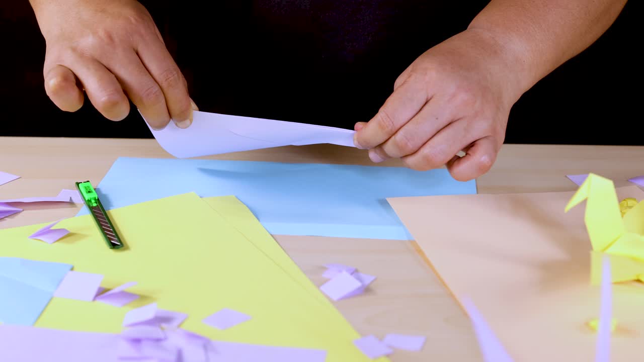 Person folds blue origami paper on desk with colorful sheets, overhead view, bright lighting