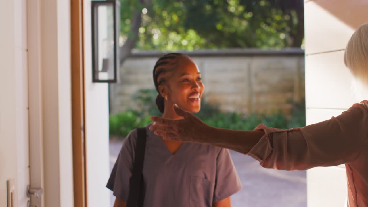 Caregiver carrying bag in scrubs reaching handle opening door hugging senior woman at home visit