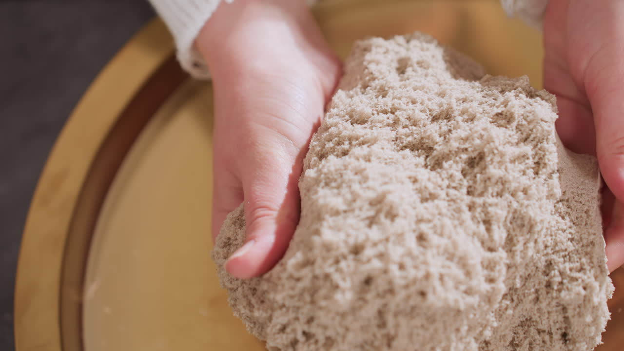High angle close up of participant gently lifting soft soil mold with both hands during calming sensory therapy session, showing textured material on golden tray in peaceful indoor environment