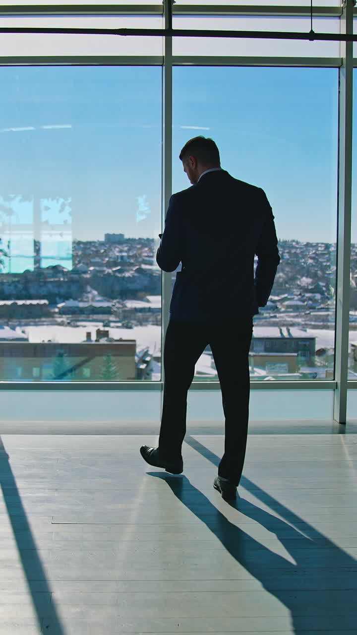 Full-length portrait of businessman entrepreneur in elegant suit walking in modern office and looking at his smartphone. Panoramic windows view on city. Vertical video