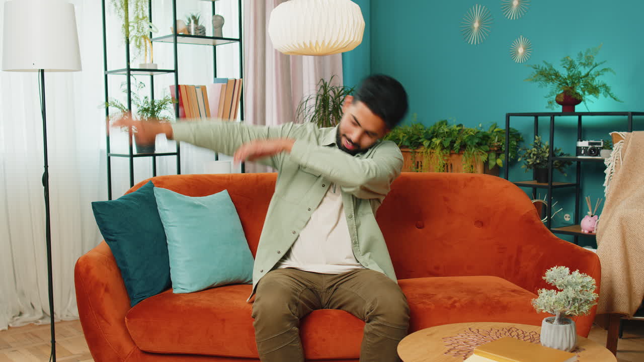 Happy man having fun, dancing, dabbing raising hands, making dub dance gesture at home room on sofa