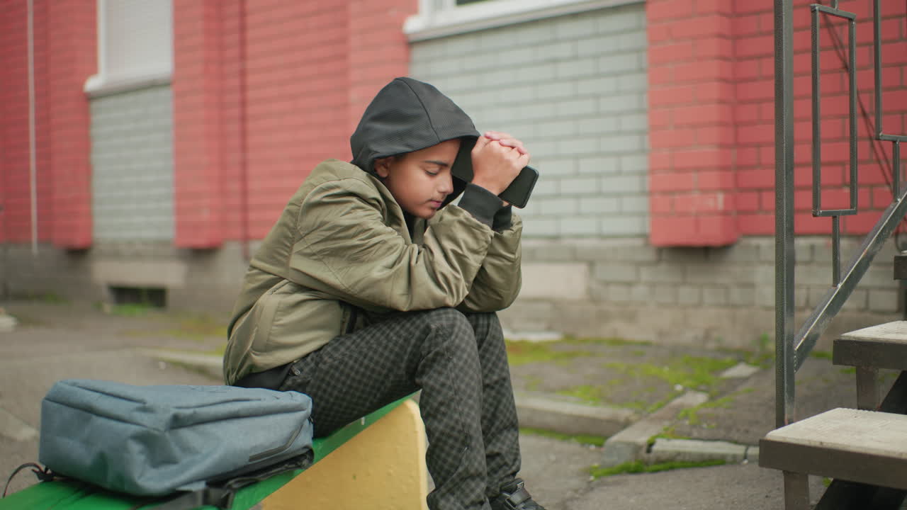 Side view of teen in hooded jacket sitting near brick building steps, holding phone with both hands and resting head gently on device, appearing thoughtful and reflective outdoors