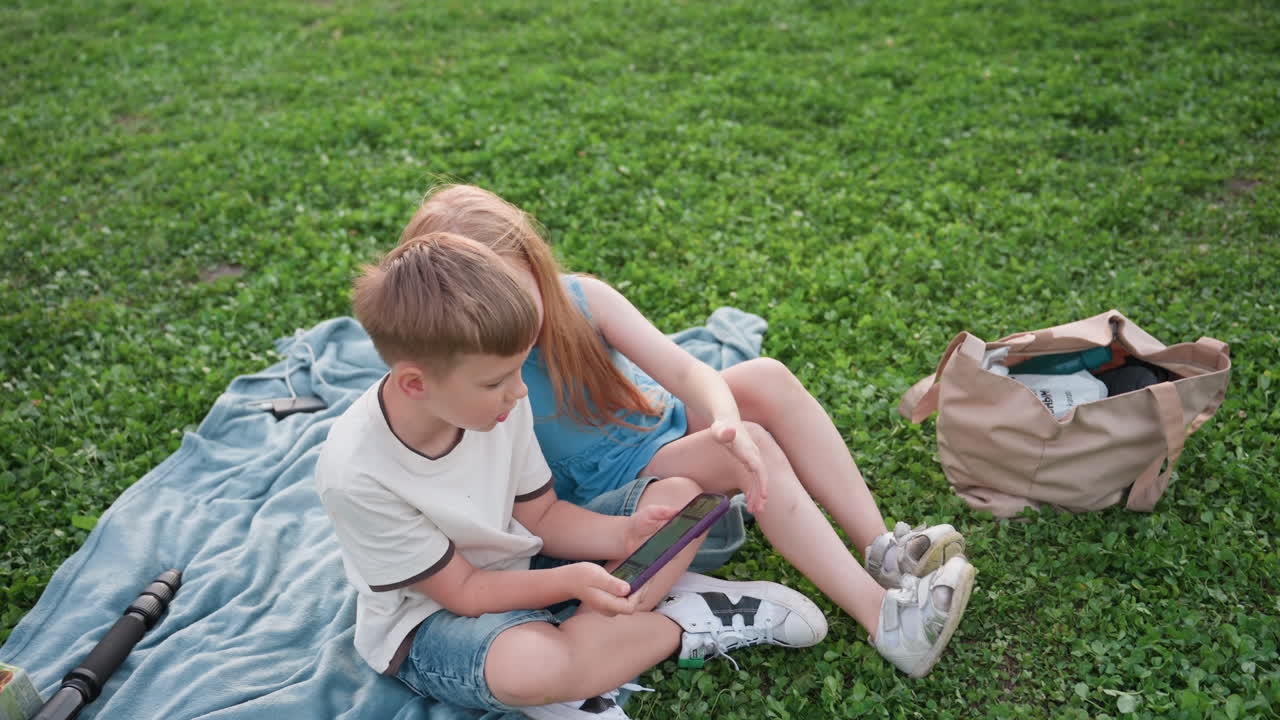 high angle view of children sitting picnic blanket green grass watching video on phone together, sibling bond, casual summer clothes, nearby bag and tripod, soft light, outdoor leisure in park