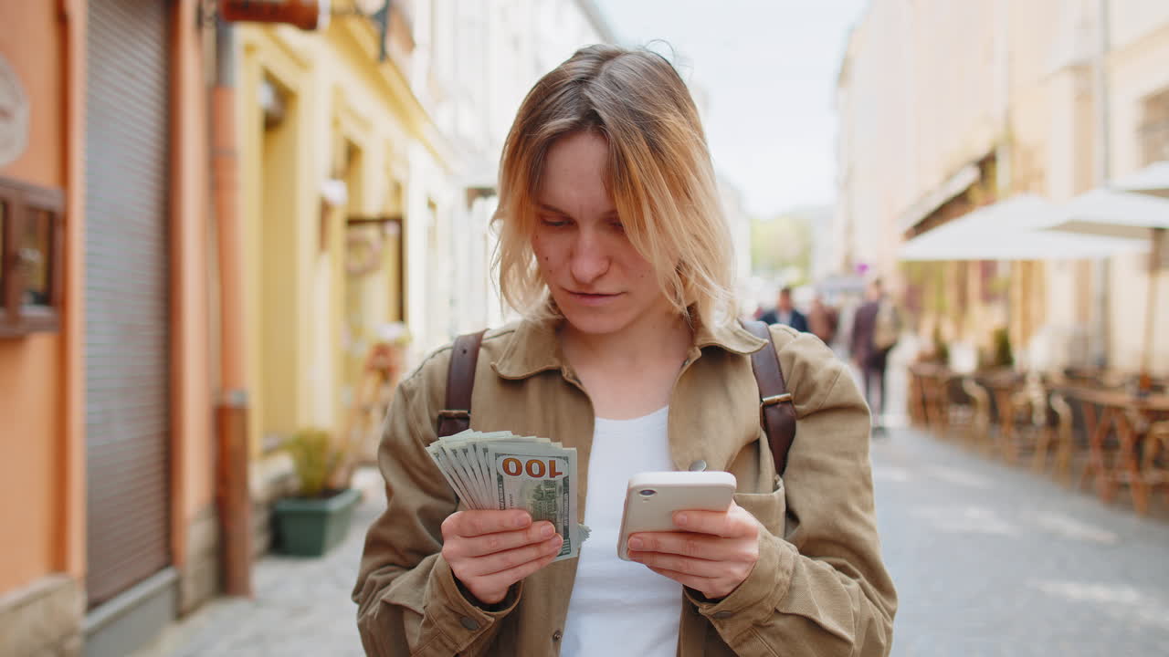 Woman counting money dollar cash using smartphone calculator app satisfied of income on city street
