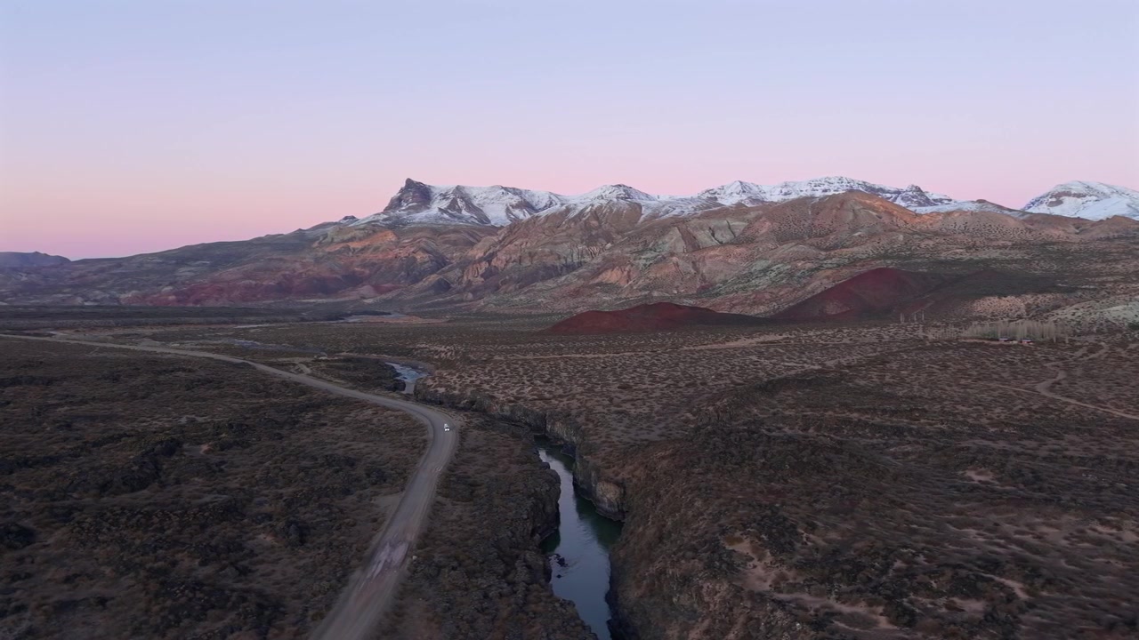 Strada sterrata solitaria che costeggia un profondo canyon vulcanico a La Payunia, con lo sfondo di cieli viola pastello all'alba. Ripresa aerea lenta, con movimento a destra.