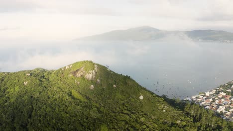 Drohne-Schwenkt-An-Einem-Sommertag-Um-Einen-Wolkigen-Gipfel-Eines-Tropischen-Waldberges-Mit-Einer-Stadt-Und-Einem-Meer-Im-Hintergrund