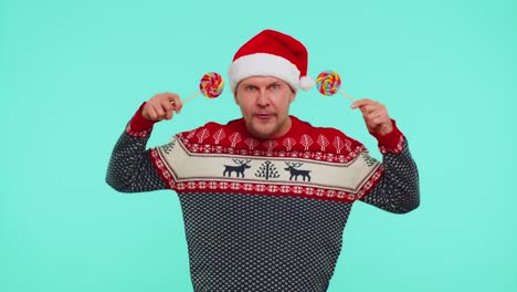 Funny-man-in-red-New-Year-sweater-holding-candy-striped-lollipops-hiding-behind-them,-fooling-around