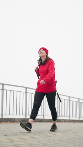 happy business woman in red sweater and head warmer with headset around neck walks casually along interlocked path holding bag over shoulder, surrounded by foggy atmosphere and calm serene