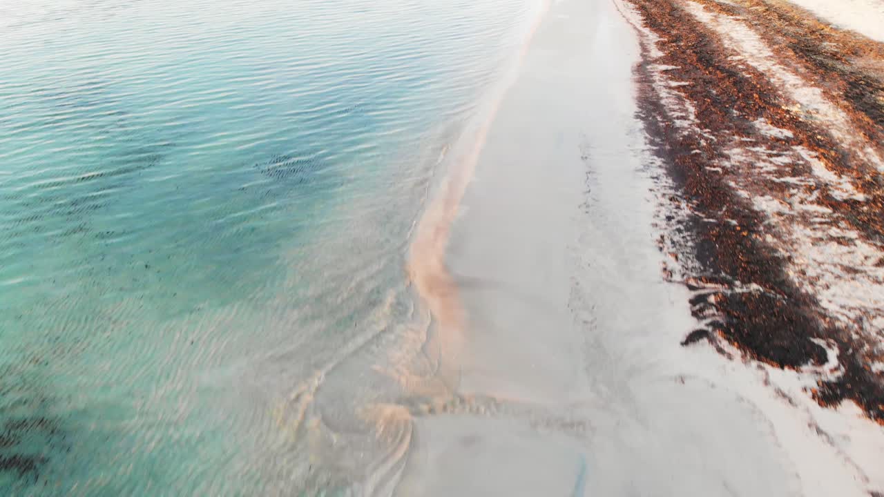 aerial drone shot panning forward and up along the waterline of blue water on a beach in Majorca at sunrise with white sand and seaweed