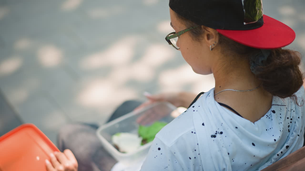 primer plano de una gorra y manos en un banco, camisa blanca moteada, visera roja, coleta y gafas de sol sobre la gorra, sombras moteadas de los árboles, reloj en la muñeca, postura relajada, recipiente para el almuerzo a un lado, tranquilidad