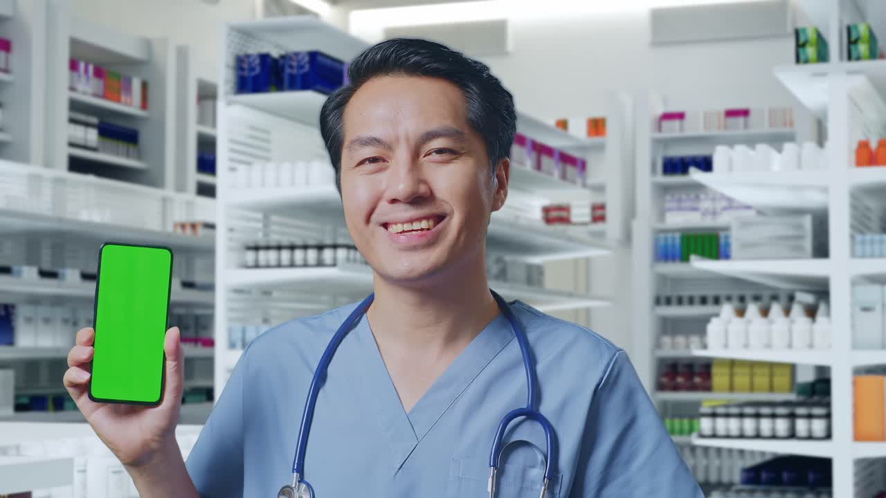 스테토스코프를 들고 있는 아시아 남성 의사가 약국에 서서 미소 짓고 녹색 화면의 스마트폰을 보여준다.