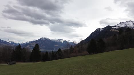 Aerial-view-of-drone-flying-through-grass-field-with-snow-capped-mountains-in-background-on-a-cloudy-day-in-Vorarlberg,-Austria