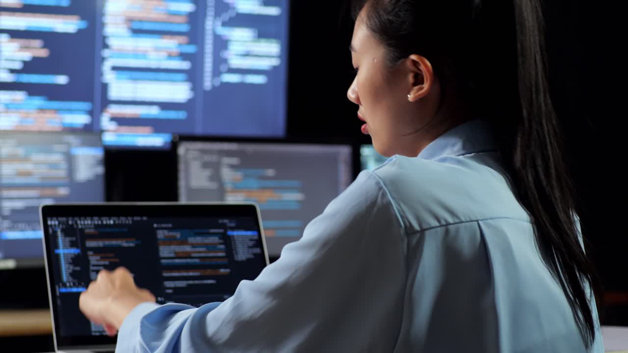vista de cerca de atrás de una programadora asiática bostezando mientras escribe código por una computadora portátil usando múltiples monitores que muestran la base de datos en los escritorios de ventana de terminal en la oficina