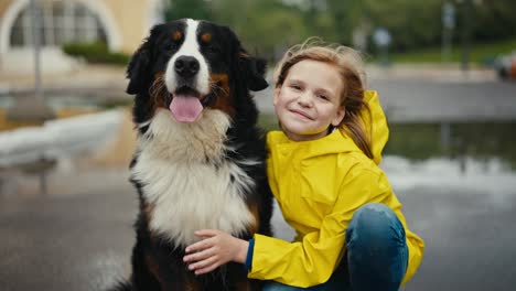 Happy-teenage-girl-in-a-yellow-jacket-posing-with-her-large-black-and-white-purebred-dog-in-the-park-after-the-rain-during-a-walk