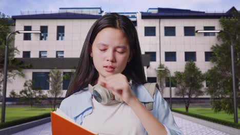 primer plano de una estudiante adolescente asiática con una mochila leyendo un libro y bostezando mientras está de pie frente a un edificio escolar