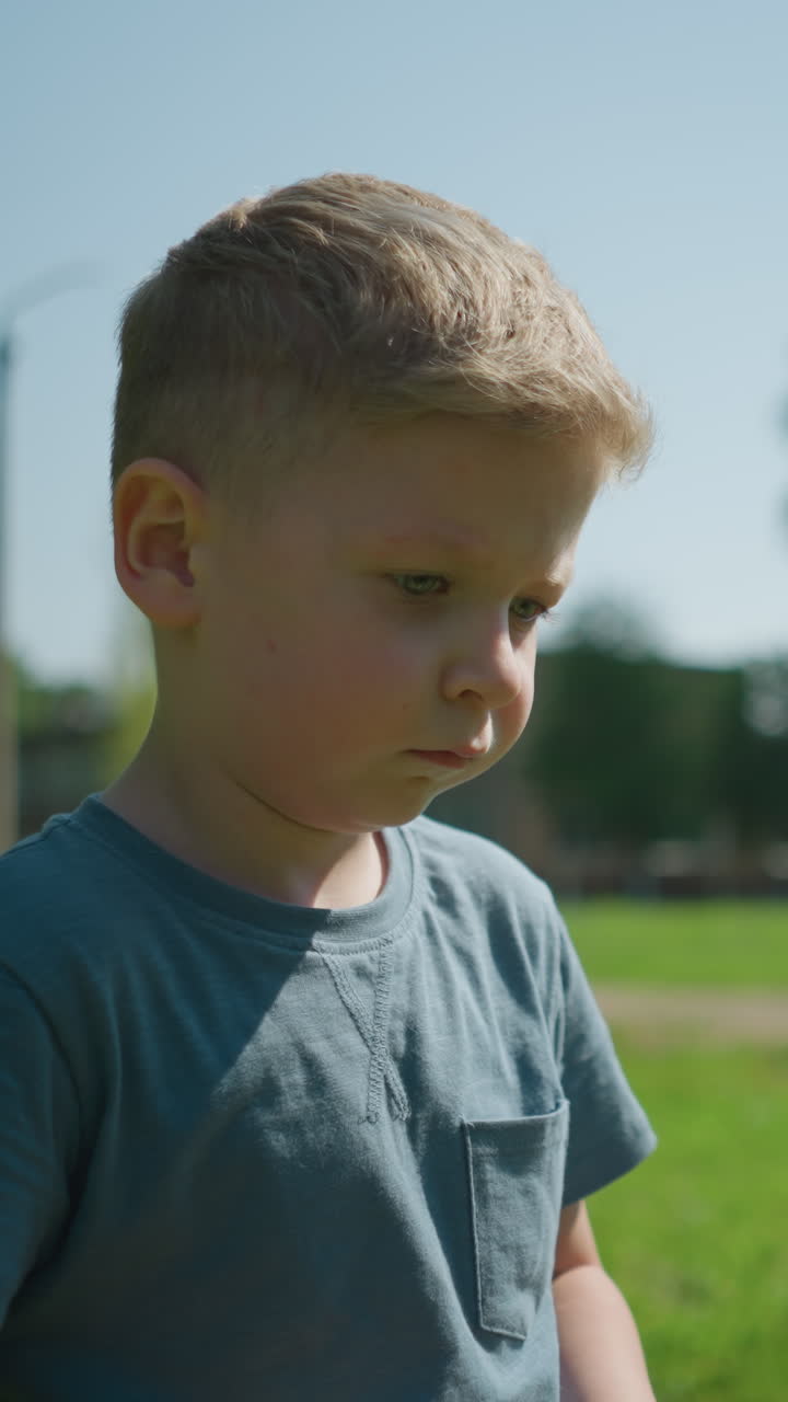 A close-up of a little child looking unhappy, with a blurred view of a woman standing in front of a goal post, set against a background of trees and a paved road in a sunny park