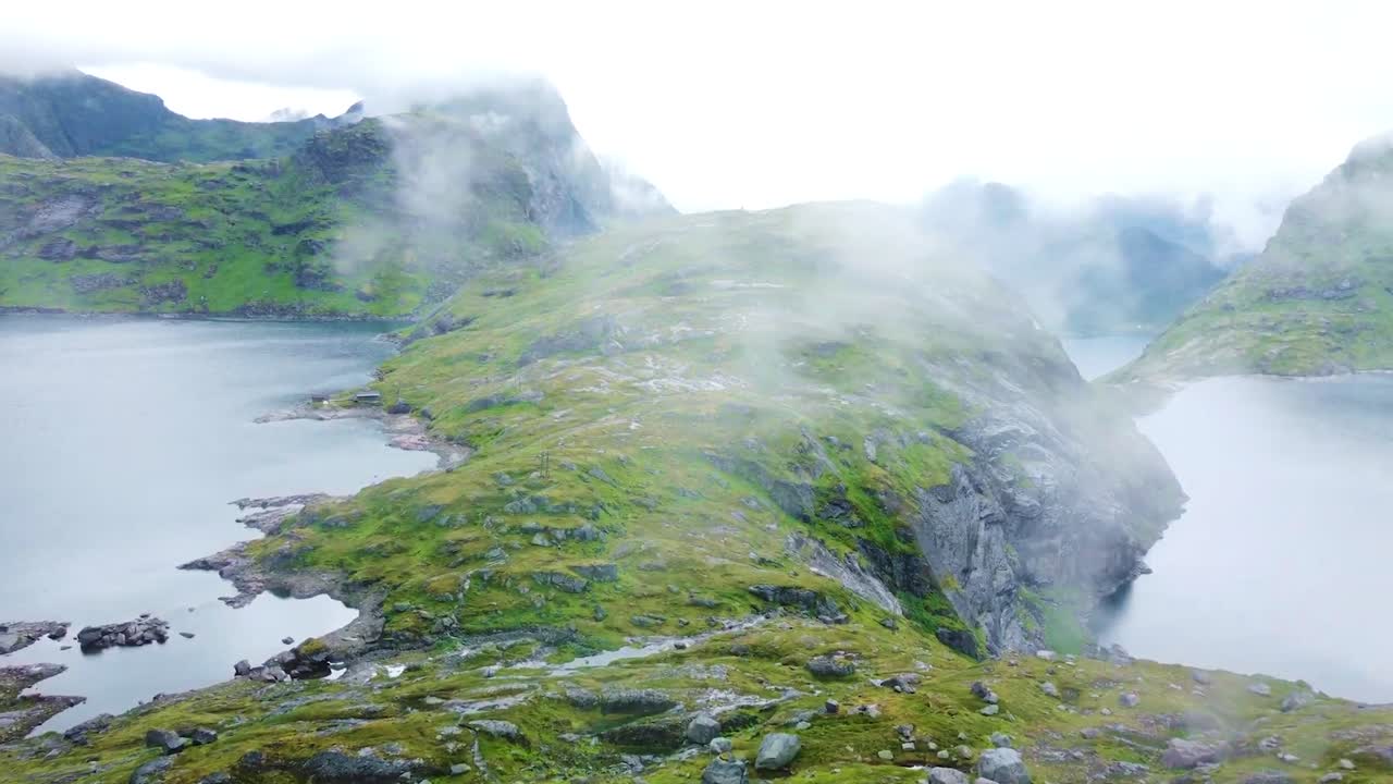 Aerial drone footage gliding and flying over Lofoten Norway rocky steep and harsh green mossy terrain and landscape with mountains with lakes visible, while fog, mist and clouds are moving on top.