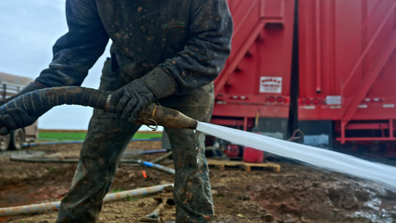 Unrecognized man in dirty clothes holding a hose with water flowing from it. Two red trucks at backdrop. Oil or gas production.