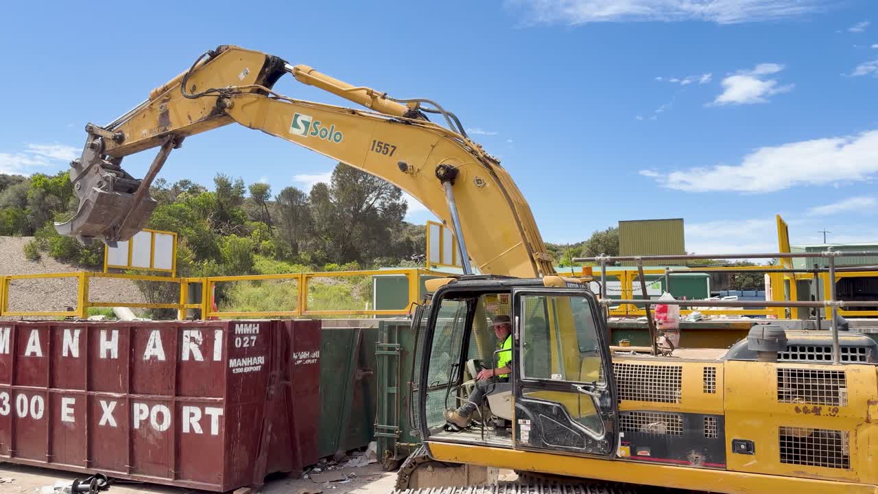 Yellow excavator transfers scrap metal into large recycling skips at outdoor waste facility under sunlight