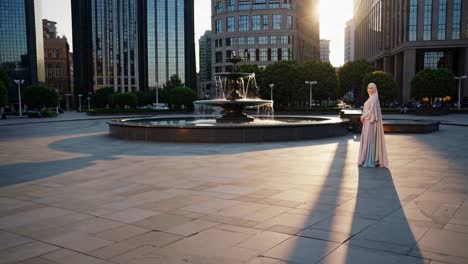 elegant woman wearing a hijab and a long dress is posing near a fountain in a modern city square, surrounded by skyscrapers and trees, with the warm sunset light creating long shadows