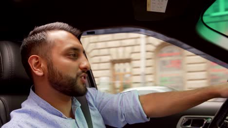 tilt up side view of young middle eastern man rapping or singing to music and dancing while driving car down city road with open window