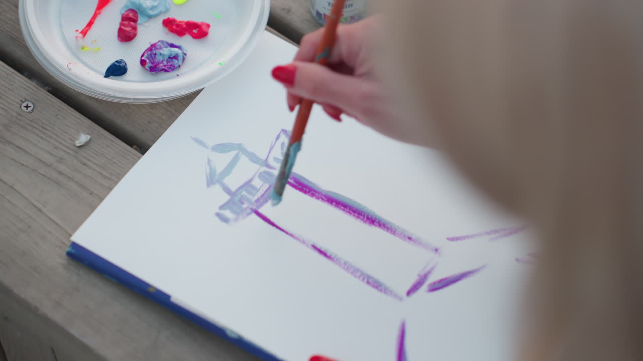 serene close up hand painting abstract tower outline with purple and blue watercolor strokes on white sheet beside colorful palette on wooden bench during sunset art session creative process