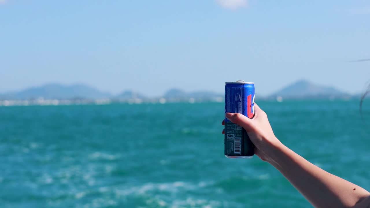 A woman shakes a cola can on a boat in Phuket, causing it to fizz over. Bright daylight and ocean backdrop