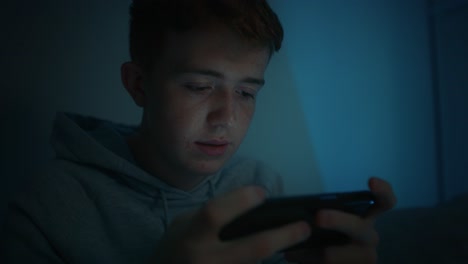 Serious-caucasian-teenage-boy-using-mobile-phone-while-sitting-at-night-in-his-room