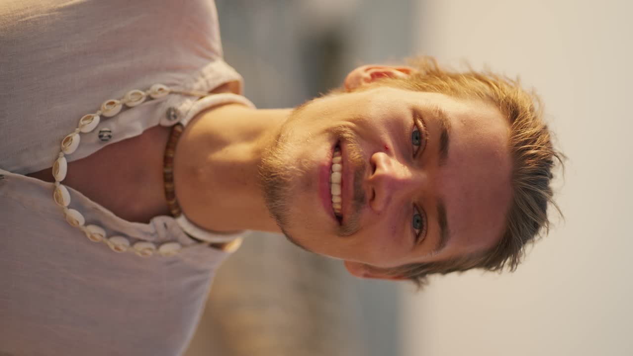 Vertical video portrait of a happy blond guy with blue eyes with stubble in a white shirt with a white necklace who stands and looks at the camera against of the sea