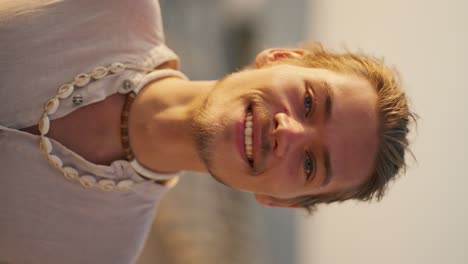 Vertical-video-portrait-of-a-happy-blond-guy-with-blue-eyes-with-stubble-in-a-white-shirt-with-a-white-necklace-who-stands-and-looks-at-the-camera-against-of-the-sea