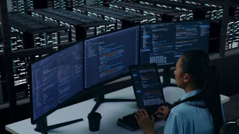 vista lateral de una desarrolladora asiática trabajando con una tableta mientras escribe código con múltiples pantallas de computadora en un centro de datos