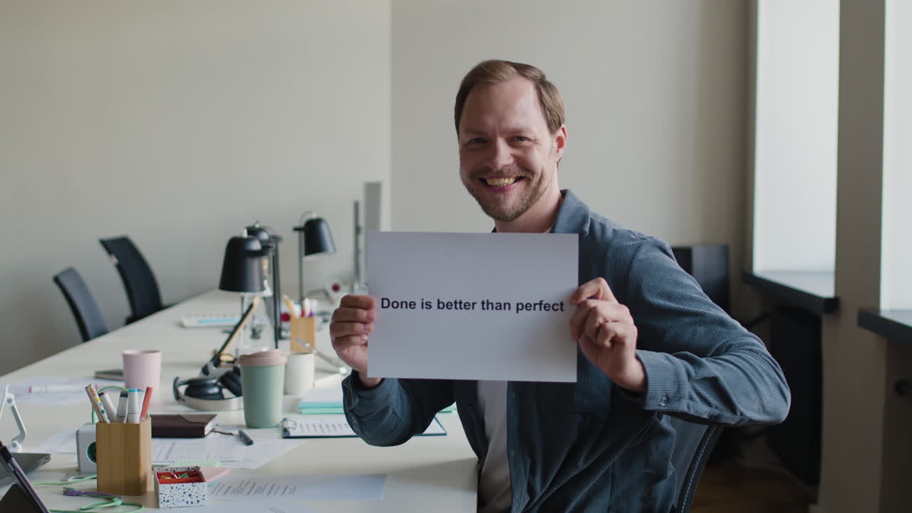 Man in an office holding a 'Done is better than perfect' sign and giving a thumbs up