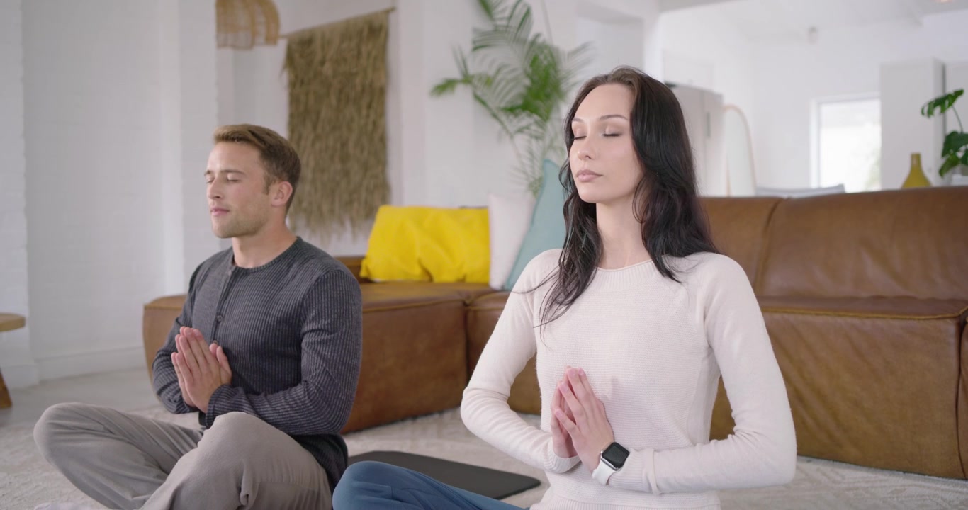 Pareja acomodándose en esterillas de yoga, en posición de oración, respirando y meditando en la sala de estar para relajarse.