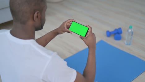vista trasera de un hombre africano mirando un teléfono inteligente con pantalla de tecla cromada en una alfombra de yoga
