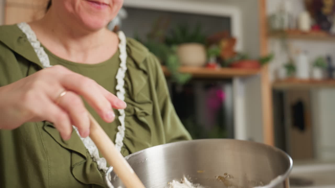 Closeup of baker in green outfit using wooden spatula to scrape thick dough from metal mixing bowl in warm home kitchen with blurred plants and shelves in background during baking process