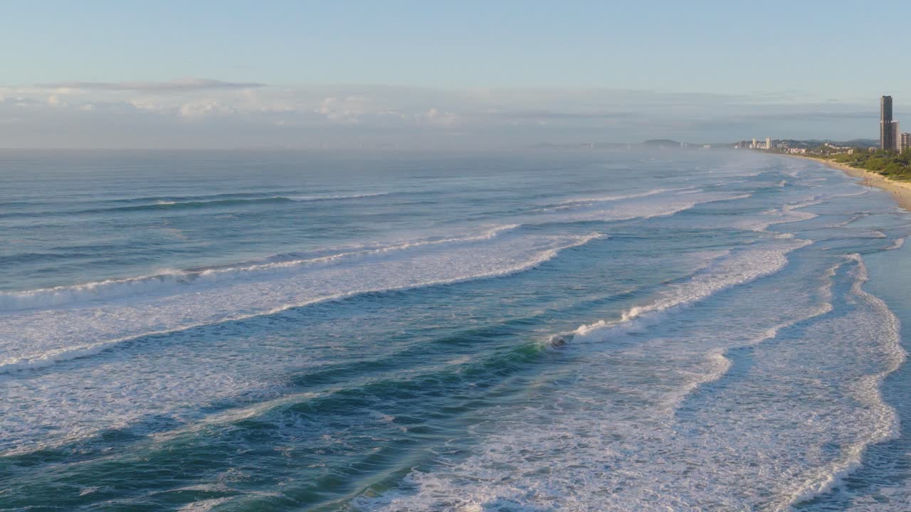Aerial footage captures waves rolling onto a serene Gold Coast beach under soft morning light, with distant buildings visible