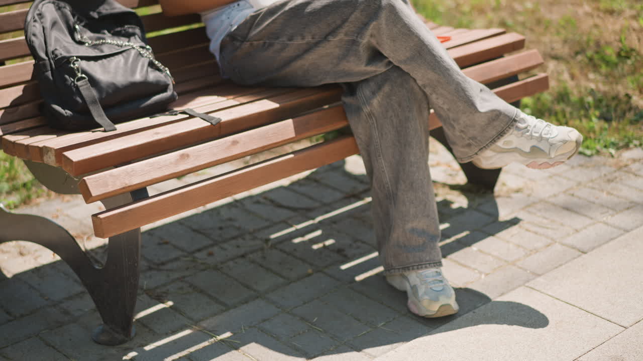 벤치, 백팩, 다리를 꼬고 앉아 햇빛 아래에서 기다리는 흰색 상의를 입은 학생, 캐주얼한 바지와 스니커즈가 보임, 잔디와 보도가 있는 도시 공원 배경, 약간의 초조함과 움직임이 느껴지는 모습