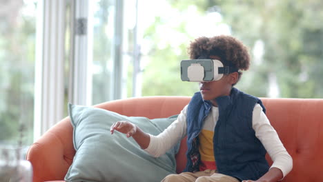 african american boy sitting on sofa, using vr headset and touching virtual screen, slow motion