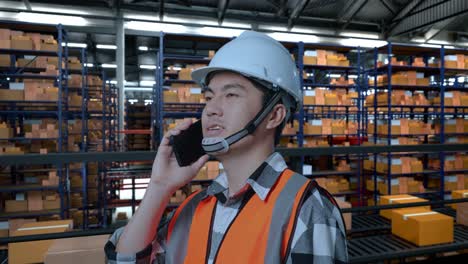 vista lateral de cerca de un ingeniero masculino asiático con casco de seguridad hablando en teléfono inteligente mientras está de pie en el almacén con estantes llenos de productos de entrega