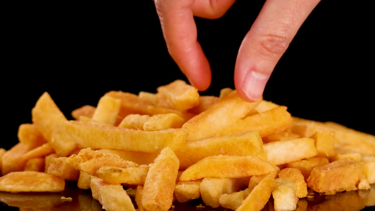 Human hand selects golden fried French fry from pile, close-up, dramatic studio lighting, static shot