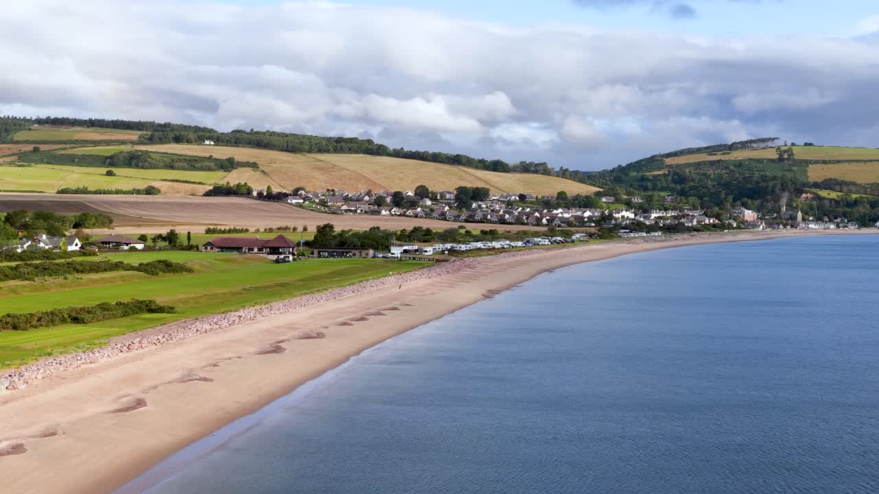 Drone camera smoothly pans along tranquil shoreline, revealing sandy beach, village, and rolling hills