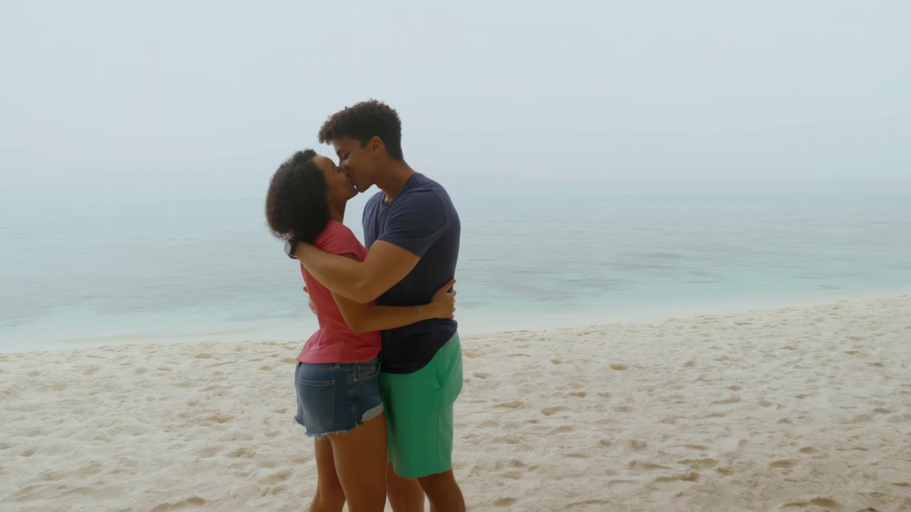 Young man and woman hold each other closely on pale sand beside calm shoreline under soft overcast light, barefoot and turned inward, creating quiet coastal mood with gentle affection