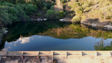 reverse flight with a drone filming a small water dam in a forest area where initially we see the striking reflection of the vegetation in the water and as we move away we see the surroundings