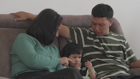 Young-Asian-Parents-Teaching-Their-Daughter-How-To-Using-Smartphone-While-Sitting-On-Sofa-Together-At-Home