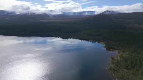 Antena-Que-Se-Eleva-Desde-Loch-Morlich,-Montañas-Nevadas-En-El-Fondo,-Escocia