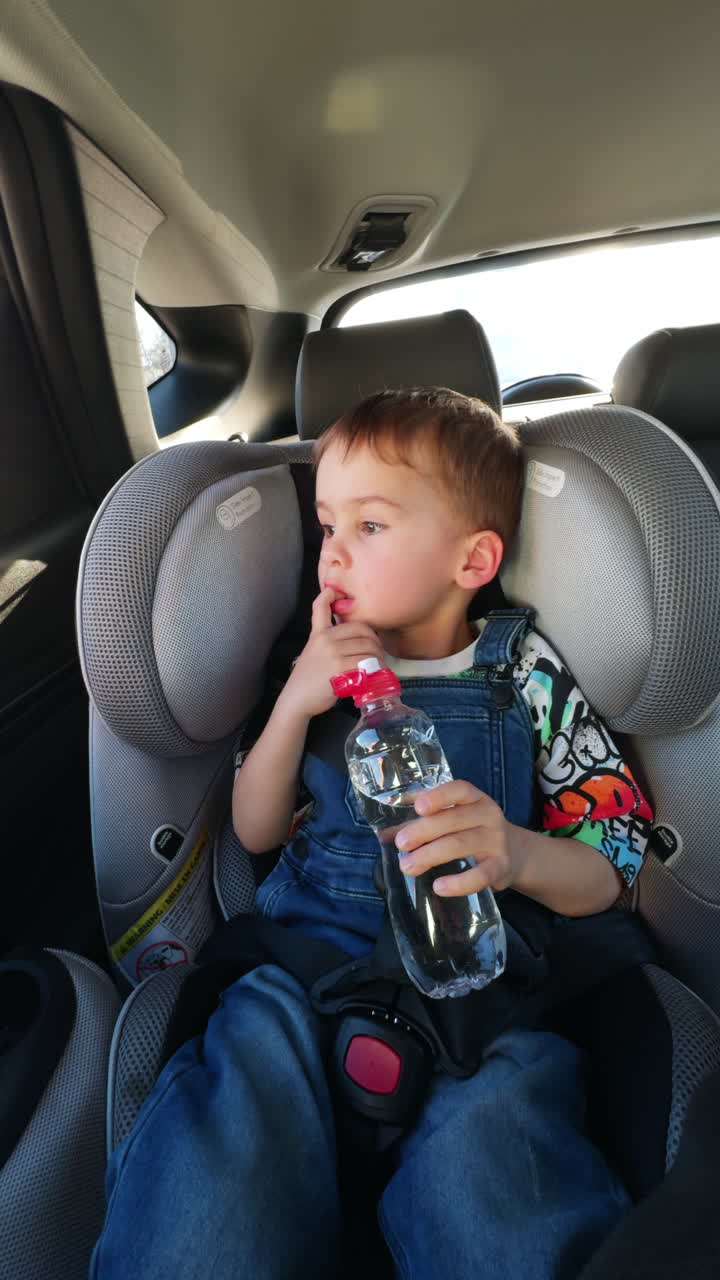 Beautiful baby boy sitting in car seat drinking water from a bottle. Toddler kid in jeans romper looks into a window. Vertical video.