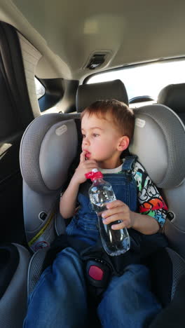 beautiful baby boy sitting in car seat drinking water from a bottle. toddler kid in jeans romper looks into a window. vertical video.