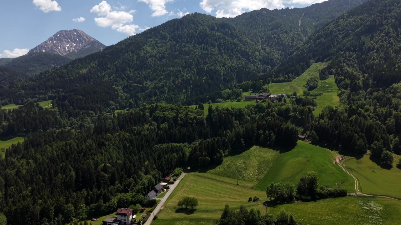 Valle alpina con prati verdi, fitta foresta, piccola fattoria, strada sterrata tortuosa, cime innevate, campagna rurale, nuvole sparse, foschia estiva, Austria, Europa. Ampia ripresa aerea con drone.
