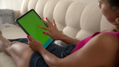 Slow-motion-of-a-mexican-latin-woman-with-a-pink-top-and-a-denim-skirt-laying-on-her-couch-sofa-browsing-her-tablet-with-her-finger-downwards