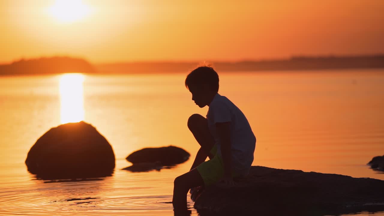 Little boy sitting on a stone among the water. Silhouette of a child near the river at a beautiful orange sunset. Slow motion.